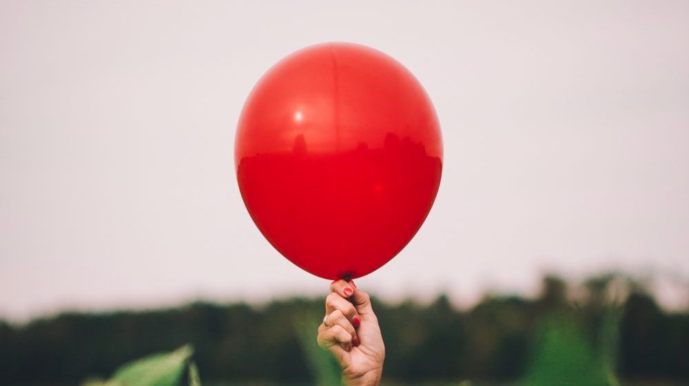 hand holding up a red balloon behind blurred plants
