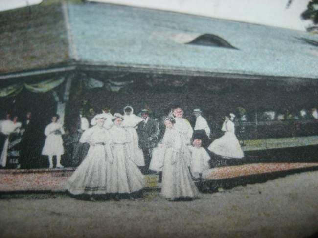 Painting of women in long white dresses with children and some men in suits outside a pavilion. 