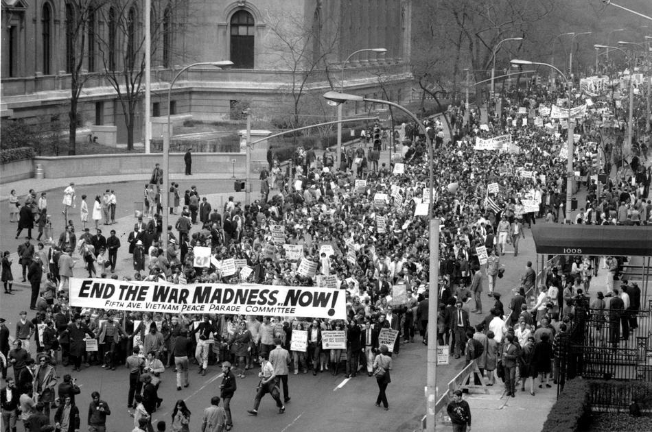 A street flooded with people protesting the Vietnam War, holding a sign that reads "end the war madness now!"