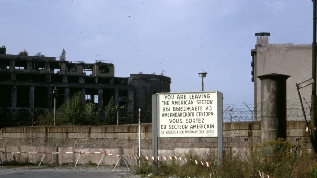 Sign reading "you are leaving the American sector" stands outside of Checkpoint Charlie, with abandoned industrial buildings in the background.
